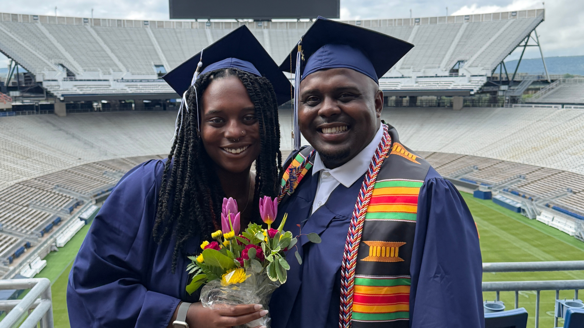 Chiara Harris (left) and Fred Quinn, alumni network coordinator at the Readiness Institute, pose together in graduation caps and gowns at Beaver Stadium during Penn State commencement ceremonies. Harris holds a bouquet of flowers while both smile toward the camera.