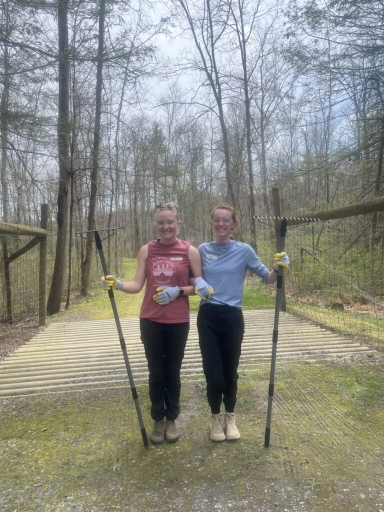 Two women pose on a bridge in the woods with rakes.