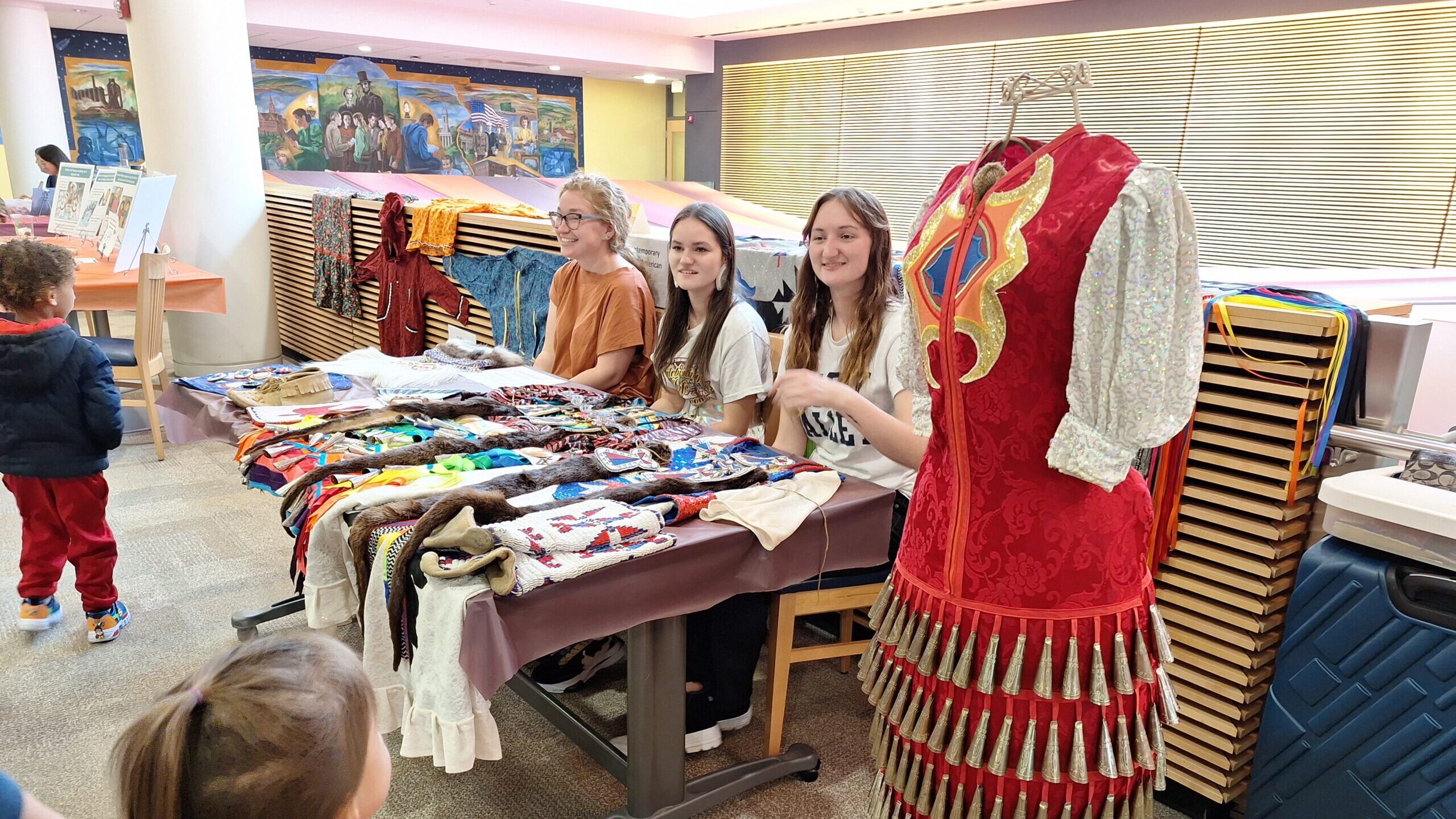 Children sit behind a table displaying colorful replicas of Native American dress.