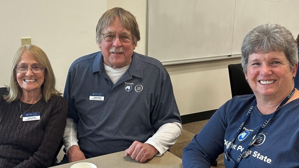 Three smiling adults sit at a table in a classroom setting, wearing Penn State OLLI name tags and shirts. In front of them are a coffee cup, a bowl with a spoon, and napkins. A whiteboard is visible in the background.