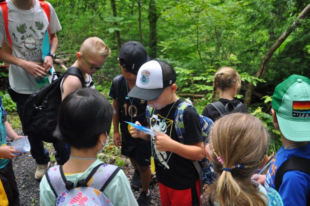 A group of young campers gathers as one of them inspects a clue on a piece of paper.