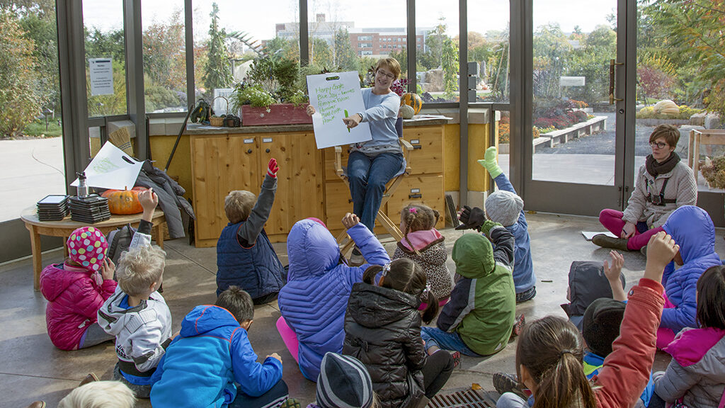 A children’s education session inside a greenhouse at the Arboretum at Penn State, with a teacher holding up a book and a group of attentive children sitting on the floor raising their hands.