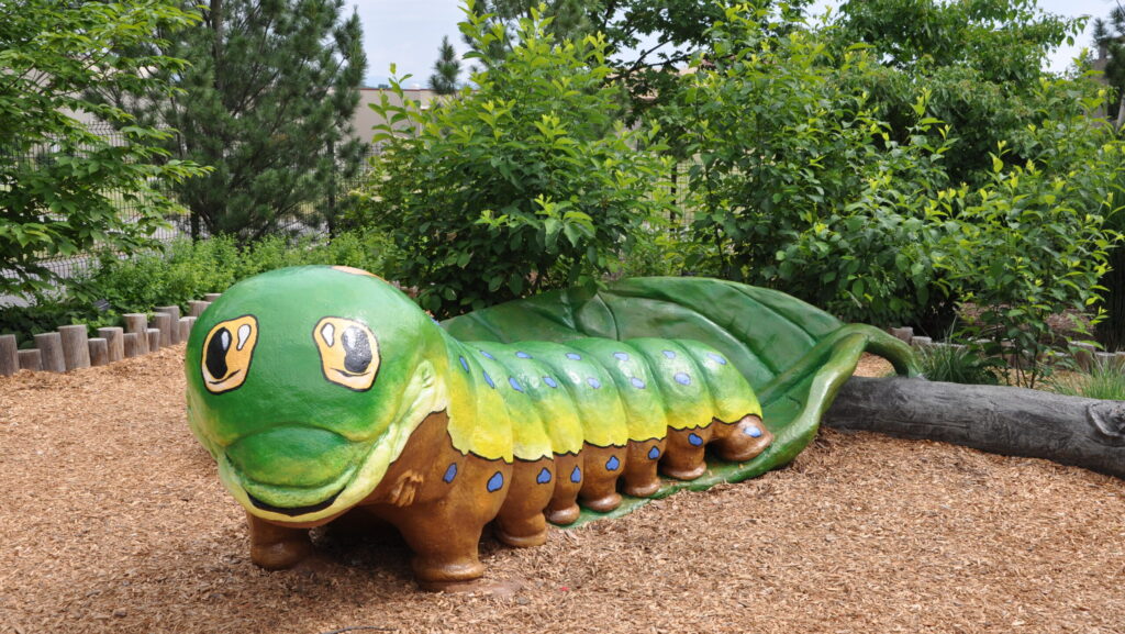 A large, colorful caterpillar sculpture at the Arboretum’s children’s garden, surrounded by wood chips, greenery, and trees in a playful outdoor space.