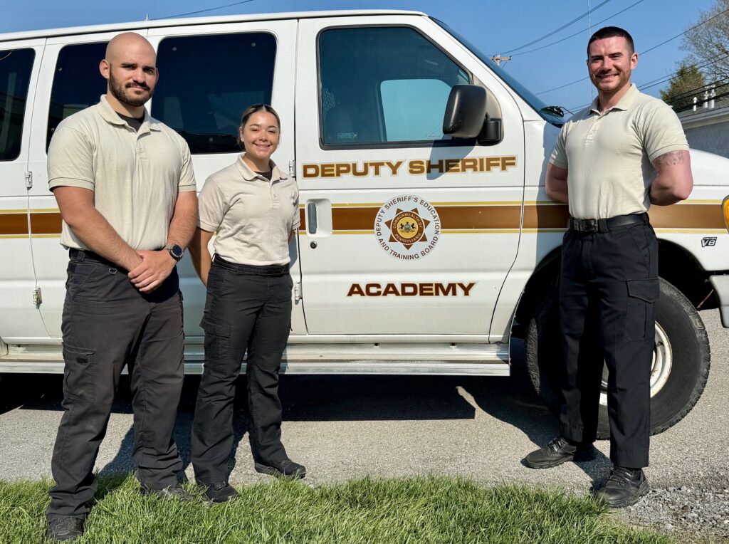 Three people dressed in uniform pose in from of a police training van.