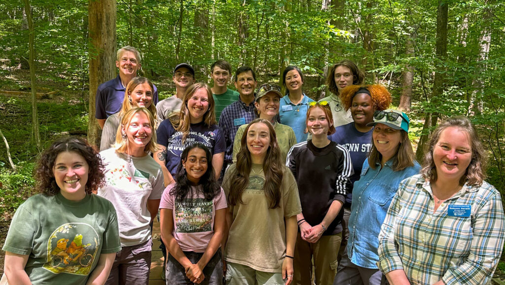 Group of students and educators posing together on a wooden platform in a lush, green forest setting, smiling as sunlight filters through the trees.
