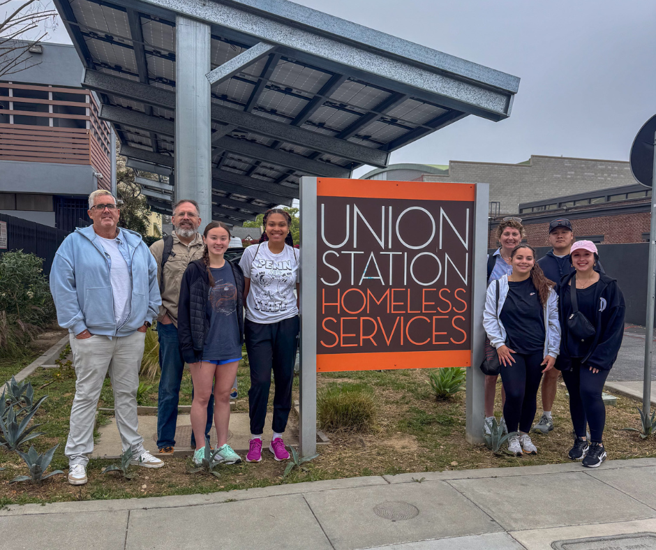 A group of nine people stand smiling in front of a sign that reads "Union Station Homeless Services." They are outside near a sidewalk with a building and solar panel canopy in the background.