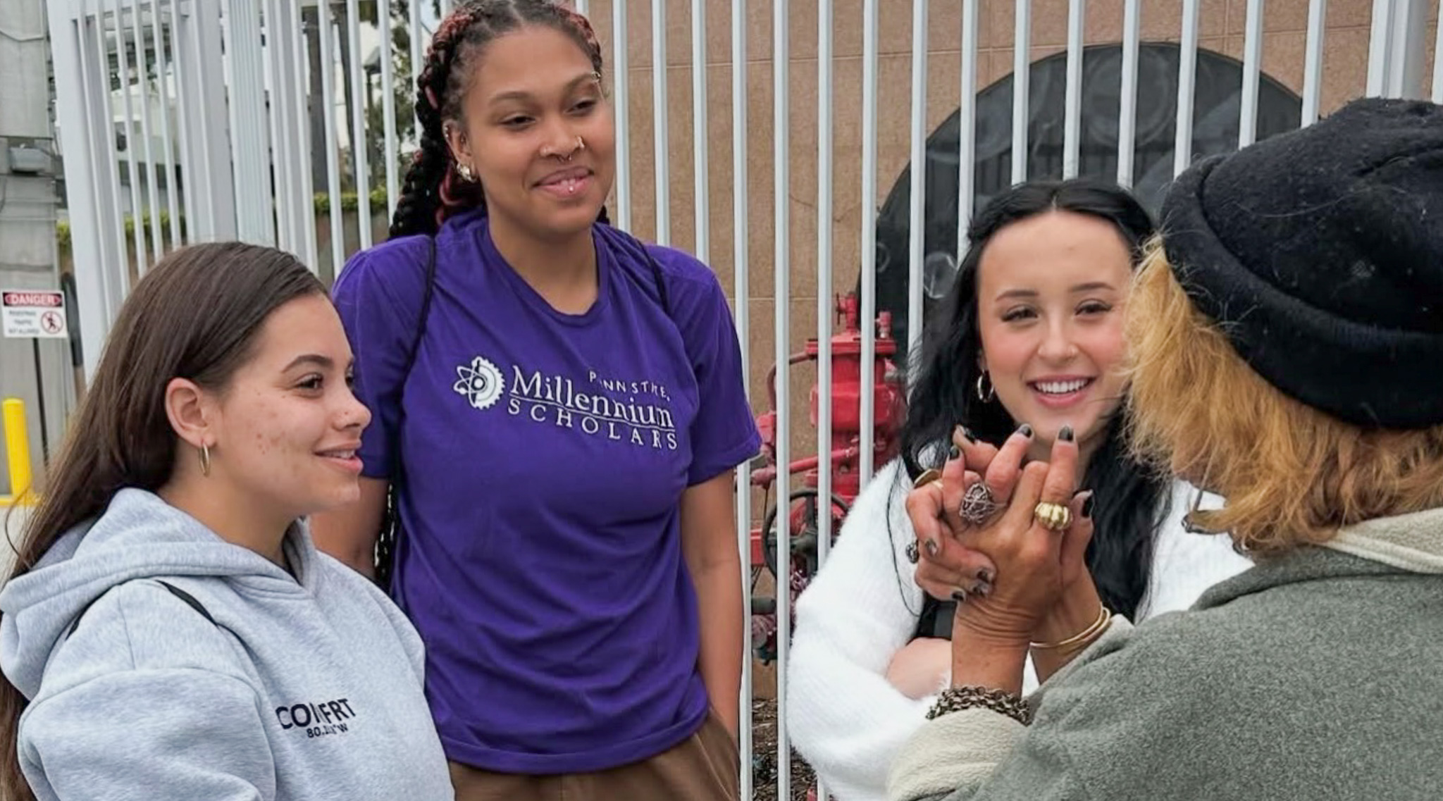 Three young women smile and listen attentively as they engage in conversation with an older woman wearing a beanie and multiple rings.