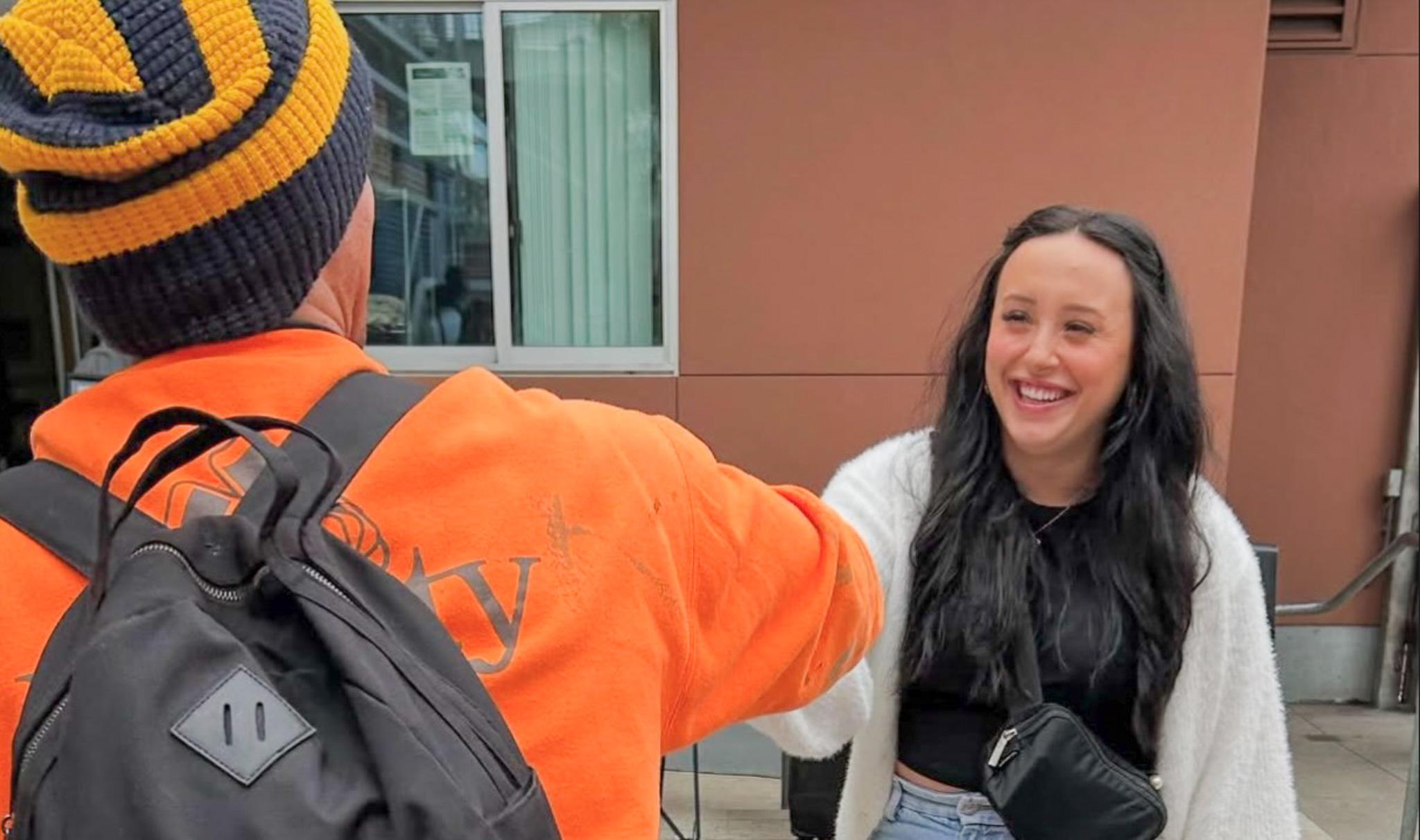 A smiling young woman with long dark hair and a white cardigan greets a man in an orange sweatshirt and striped beanie with a fist bump