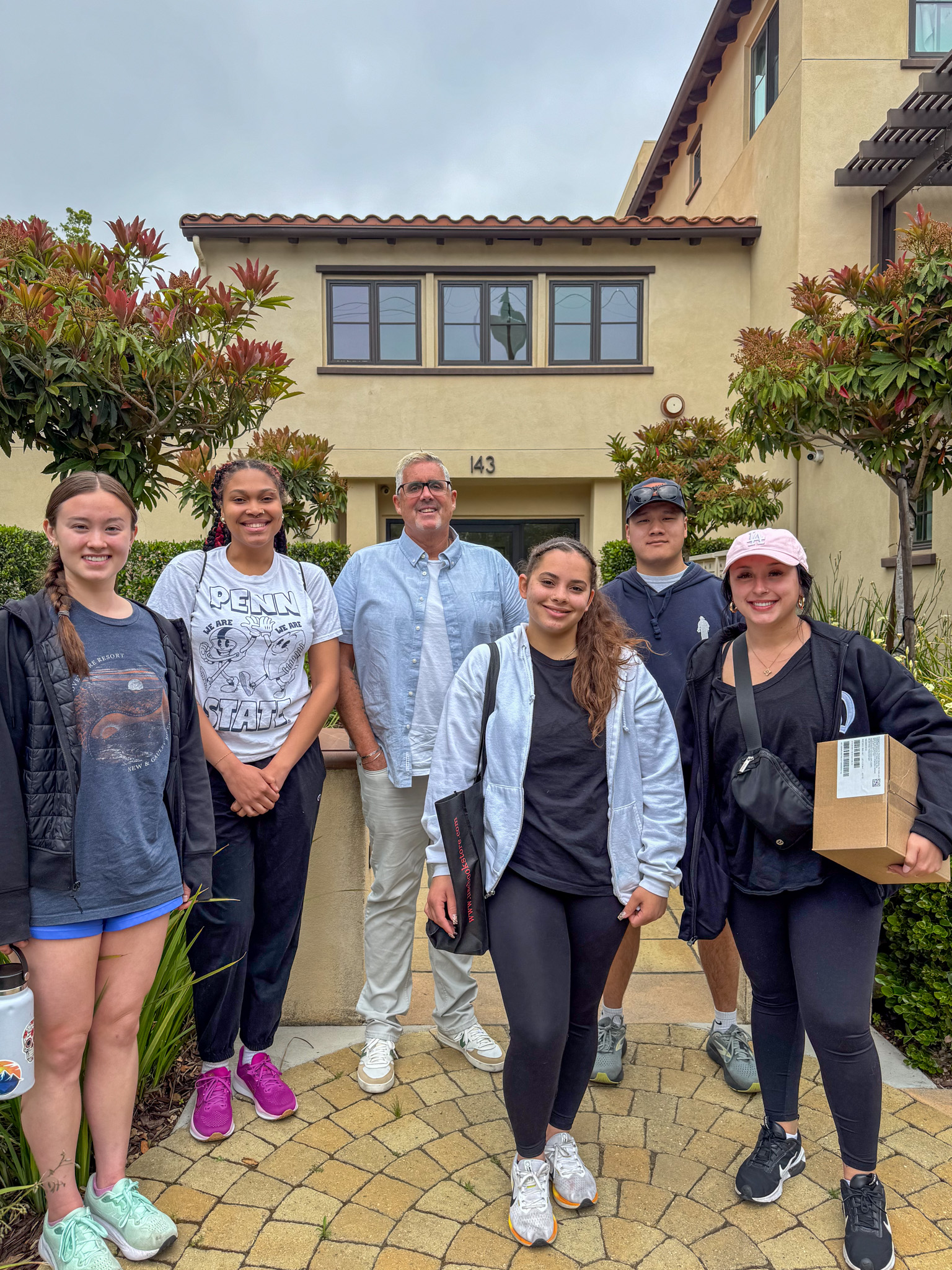 A group of six people stands smiling in front of a tan building
