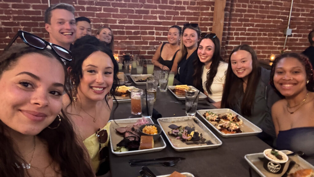 A group of smiling young adults sits around a long table at a restaurant, enjoying a meal together.