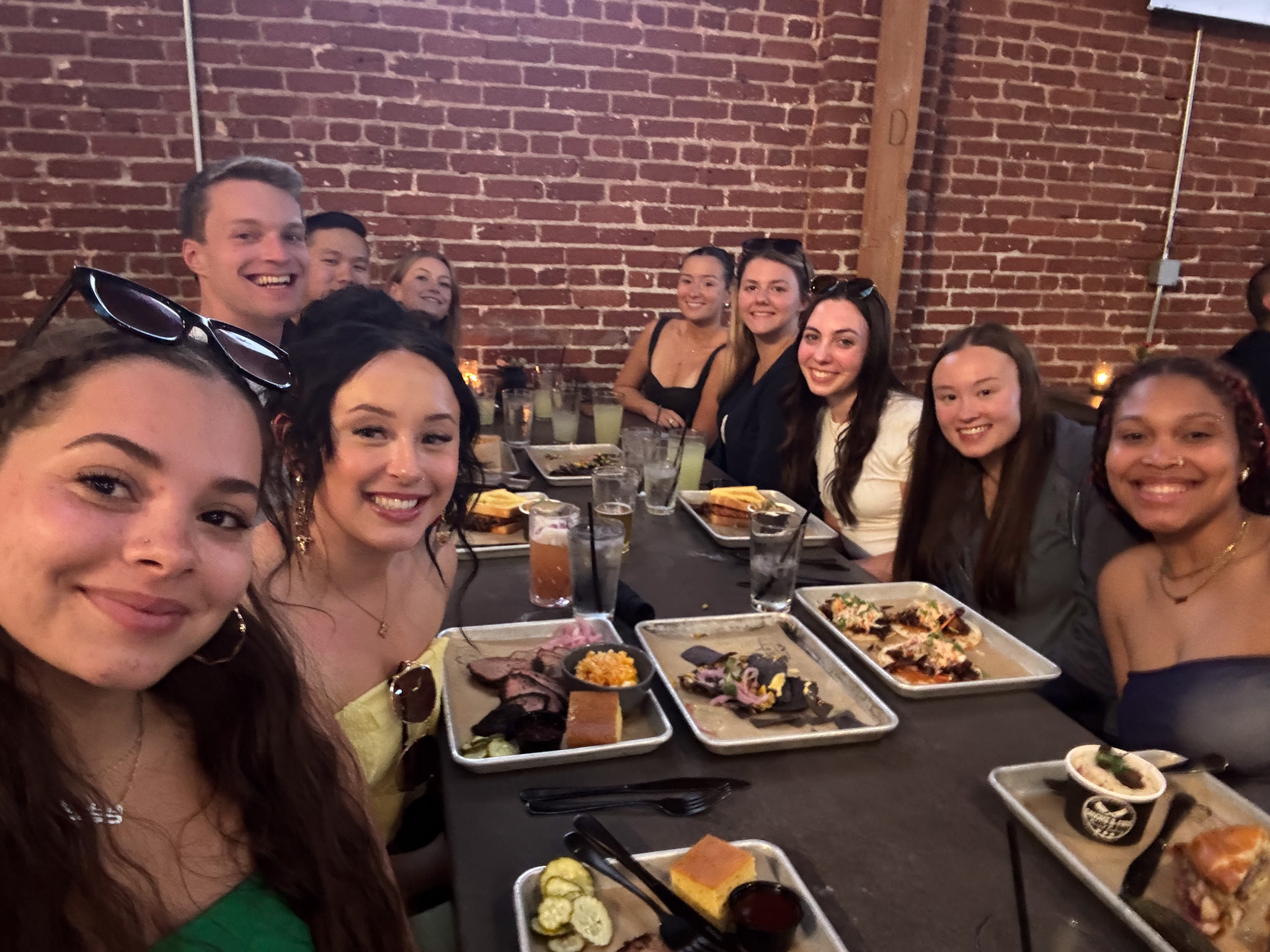 A group of smiling young adults sits around a long table at a restaurant, enjoying a meal together.